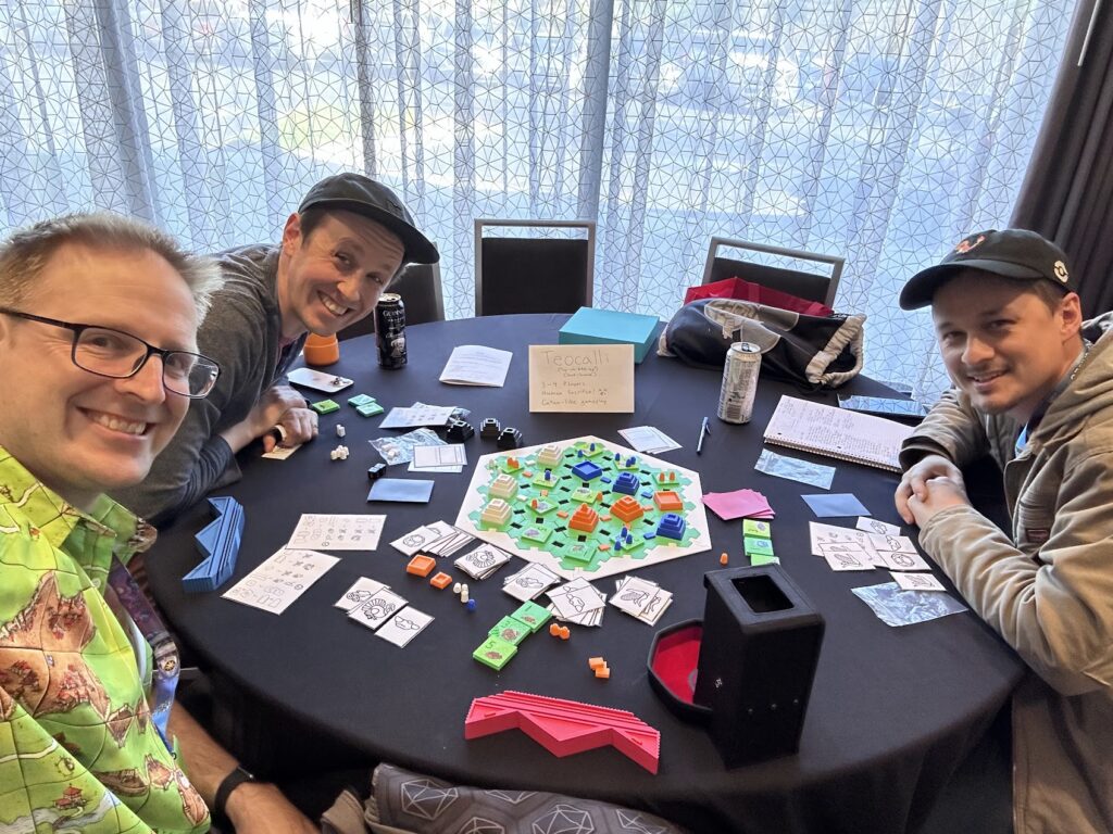 Three people seeted around a table playing the Teocalli board game.